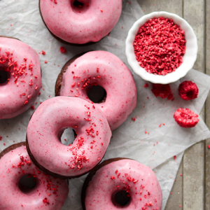 Chocolate donuts with a raspberry glaze sitting on parchment paper with freeze dried raspberries in a small bowl