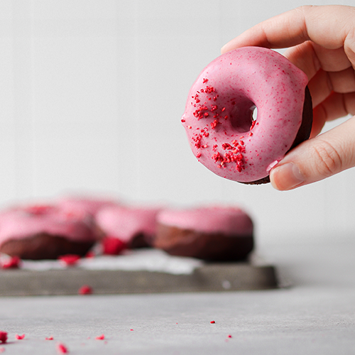 Hand holding a donut in foreground with donuts on a baking tray in the background