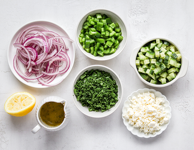 Greek salad ingredients in small bowls. These are red onion, green pepper, parsley, feta, and cucumber.